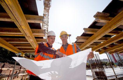 Structural engineer and architect dressed in shirts, orange work vests and helmets explore construction documentation on the building site near the wooden building constructions .