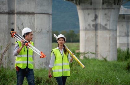 Two engineers wearing safety vests and holding surveying equipment. They are walking across a grassy field